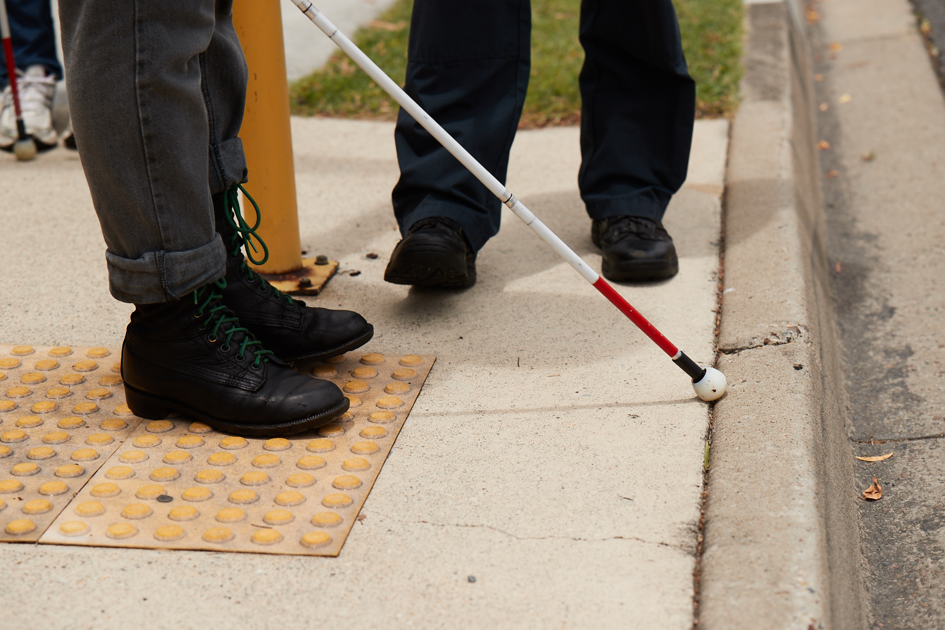 The tip of a white cane rests at the edge of a gutter