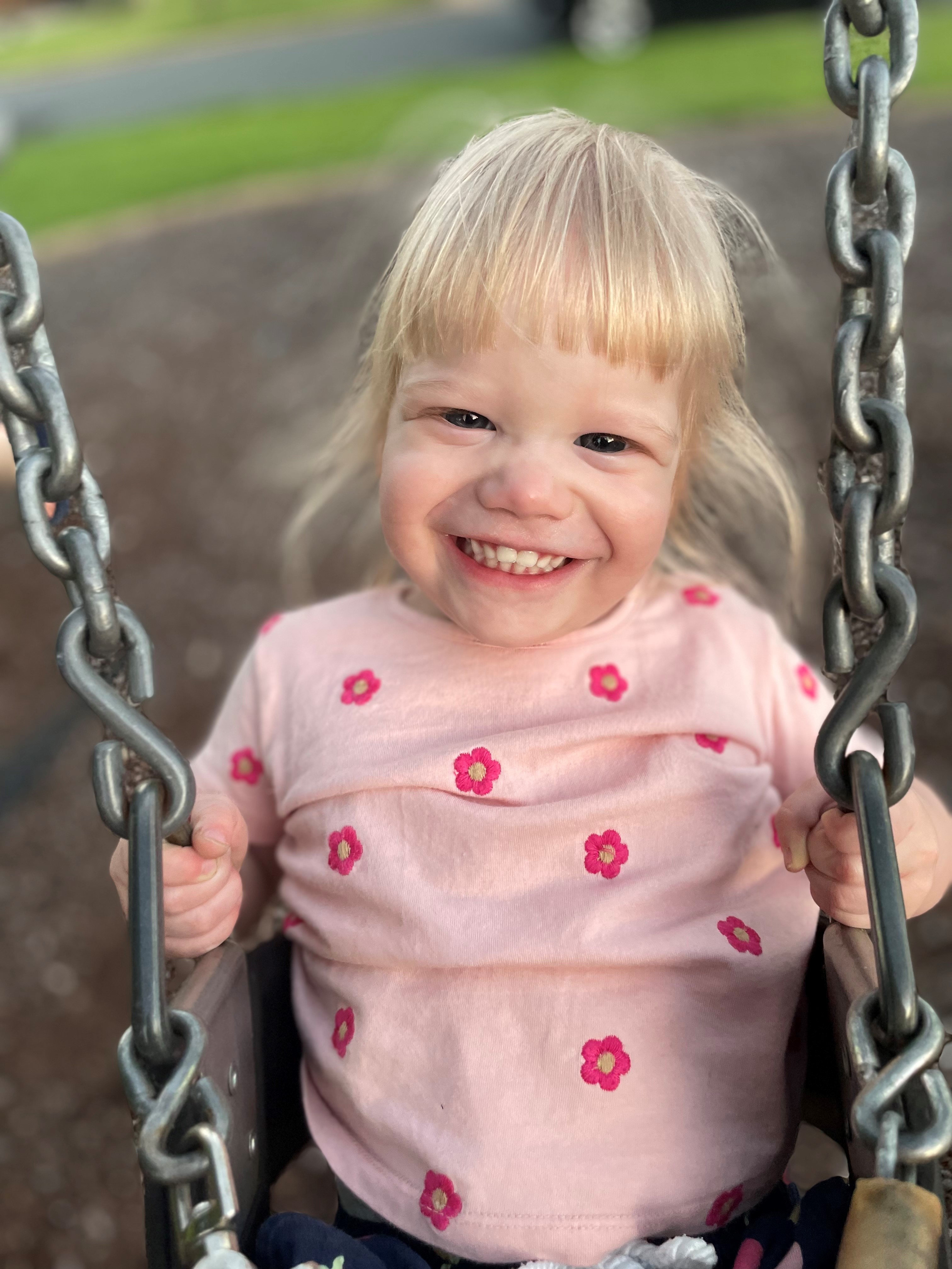 "Lucy smiling widely while sat on a swing in a playground"