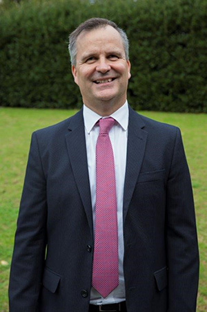 Vision Australia Manager Government Relations, Advocacy, NDIS and Aged Care Chris Edwards is smiling at the camera. He is wearing a black suit with pink tie and white shirt.