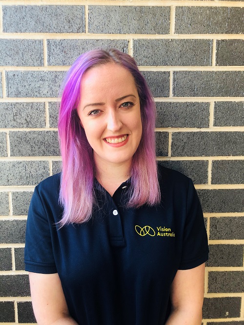 Kelsey standing front of brick wall in a Vision Australia polo shirt