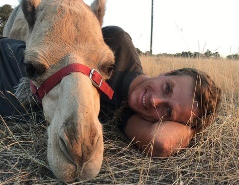 Max laying down on his side with a camel resting its head on him.