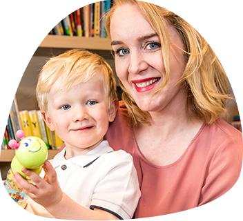 A woman with long blonde hair has her toddler son on her lap. They are both smiling at the camera.